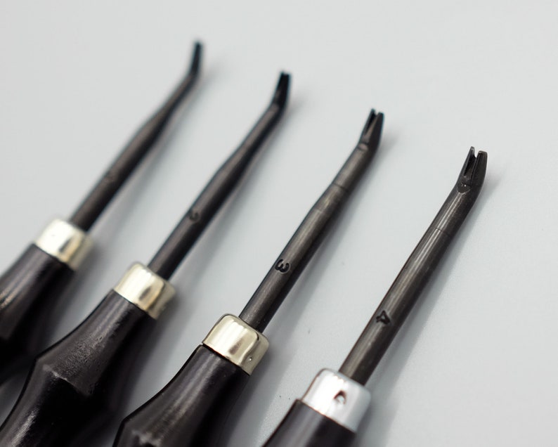 May include: Four black leather working tools with silver metal tips and numbered handles. The tools are arranged in a row on a white surface.