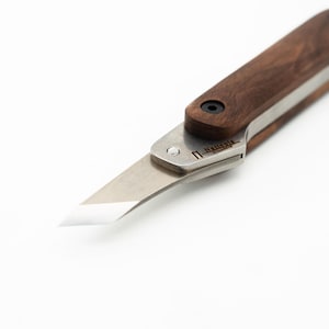 May include: A small, wooden-handled knife with a silver blade. The blade is partially extended and the handle is made of dark wood. The knife has a small silver rivet on the handle and the text "N. HALLBERG" is engraved on the blade.