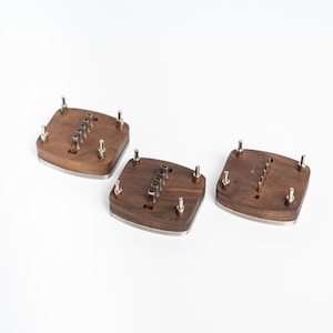 May include: Three square wooden coasters with a dark brown finish and a silver metal trim. Each coaster has a series of small metal pegs and a groove. The coasters are displayed on a white background.