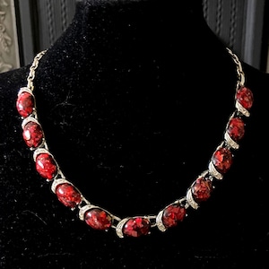 Könnte beinhalten: Eine silberfarbene Halskette mit ovalen, rot gefärbten Steinen. Die Steine sind in einem silbernen Metall mit kleinen, klaren Akzentsteinen gefasst. Die Halskette wird auf einer schwarzen Samtbüste vor dunklem Hintergrund präsentiert.