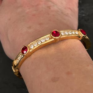 May include: Gold-tone bracelet with three red gemstones and numerous clear crystals. The bracelet is a hinged style, with a polished finish. The gemstones are round and set in a bezel setting. The clear crystals are small and pave-set.