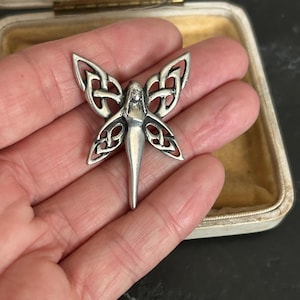 May include: A silver-toned metal brooch shaped like a fairy with wings featuring intricate openwork Celtic knot patterns. The figure has a slender body and pointed wings, with a polished finish that highlights the detailed metalwork design.
