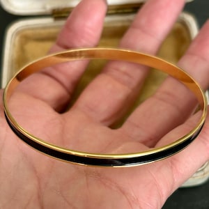 以下が含まれることがあります： 光沢のあるゴールドトーンの金属バンドに、外側の中心に沿って滑らかで光沢のある黒いエナメルのインレイが施された、細い円形のバングルブレスレット。バングルの内側は、反射性のあるゴールドトーンの仕上げになっています。