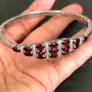 May include: A silver-toned hinged bangle bracelet featuring a central cluster of oval-cut deep red garnet gemstones. The metal band displays an intricate, openwork lattice pattern along its sides, while the stones are set in a multi-row arrangement.