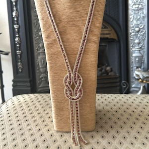 Peut inclure: Un long collier composé d'une chaîne en maille métallique dorée avec un nœud central. La chaîne est ornée d'une rangée continue de petits cristaux rouge foncé incrustés au centre, se prolongeant par deux extrémités pendantes sous le nœud.