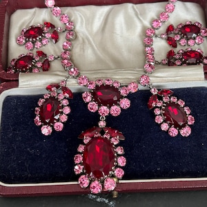 Könnte beinhalten: Eine Halskette mit einem zentralen, ovalen, tiefroten facettierten Stein, der von kleineren rosa Kristallen umgeben ist. Die Kette besteht aus rosa Kristallgliedern mit zusätzlichen Clustern aus roten und rosa Steinen an den Seiten.