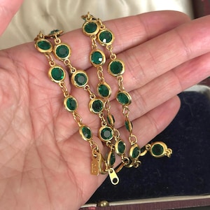 May include: A gold bracelet with emerald-green gemstones. The bracelet has a delicate chain design with each round gemstone set in a gold bezel. The clasp is visible, and the bracelet is displayed against a dark blue velvet background.