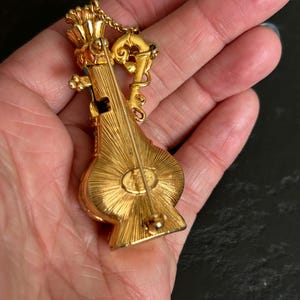 May include: A gold-toned brooch in the shape of a musical instrument, possibly a lute or mandolin. The brooch has intricate detailing, including a crown-like element at the top and a decorative chain. The back of the brooch has a pin for attachment.