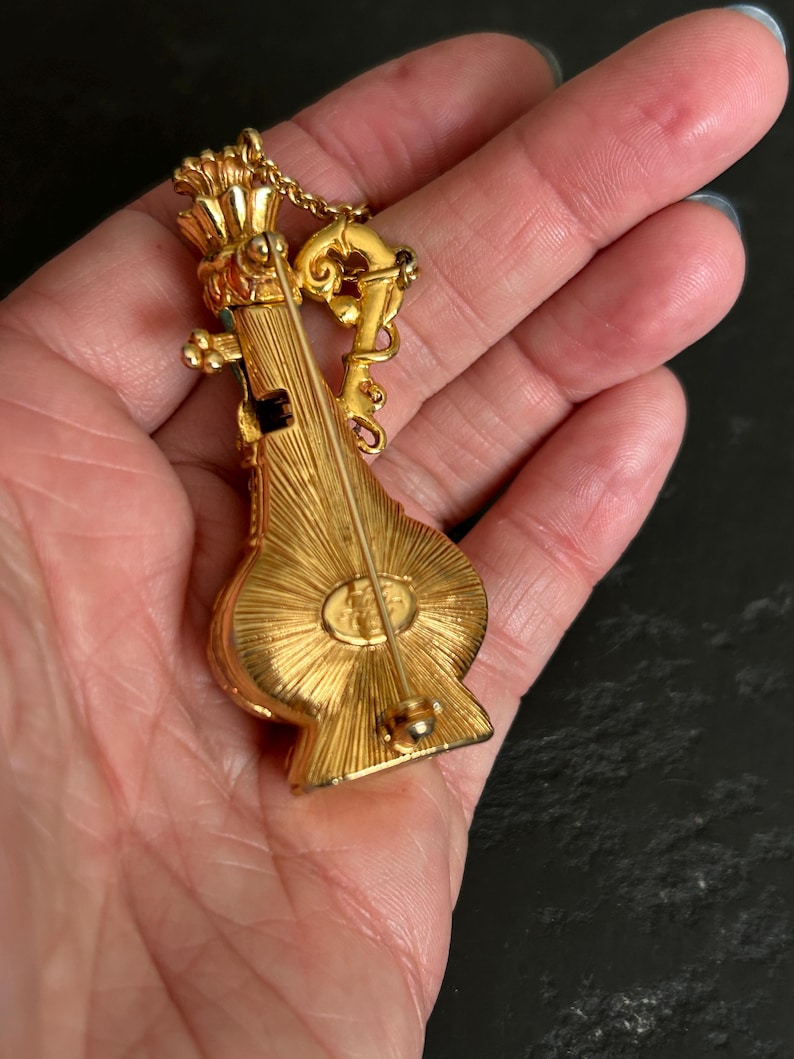 May include: A gold-toned brooch in the shape of a musical instrument, possibly a lute or mandolin. The brooch has intricate detailing, including a crown-like element at the top and a decorative chain. The back of the brooch has a pin for attachment.