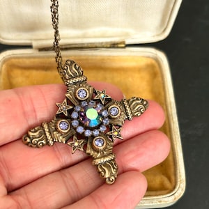 Könnte beinhalten: Eine Halskette mit einem Kreuzanhänger im Vintage-Stil in Bronze. Das Kreuz hat einen großen, schillernden Mittelstein, der von kleineren lila Steinen und Sternakzenten umgeben ist. Die Halskette wird in einer offenen Schmuckschatulle präsentiert.