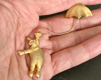 May include: A gold-toned metal brooch featuring a clown figure with a ruffled collar and three buttons on the torso. The clown is connected by a thin, twisted gold chain to a separate gold-toned umbrella-shaped piece with a pin backing.