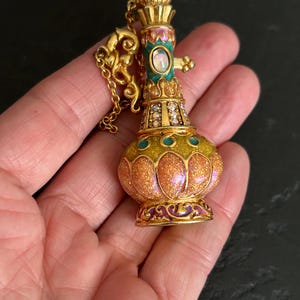 May include: A gold-colored pendant necklace in the shape of a decorative bottle. The bottle has a bulbous body with a glittery orange and yellow finish, accented with teal and purple enamel. The top is adorned with a crown-like design and a small chain.
