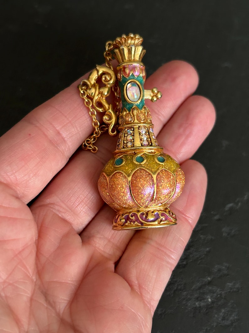 May include: A gold-colored pendant necklace in the shape of a decorative bottle. The bottle has a bulbous body with a glittery orange and yellow finish, accented with teal and purple enamel. The top is adorned with a crown-like design and a small chain.
