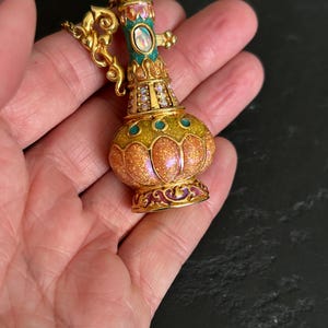 May include: A gold-toned, ornate perfume bottle pendant with a bulbous body and a slender neck. The bottle features a glittery orange and yellow body, teal accents, and a central oval-shaped gem. The pendant is embellished with small crystals and intricate gold detailing.
