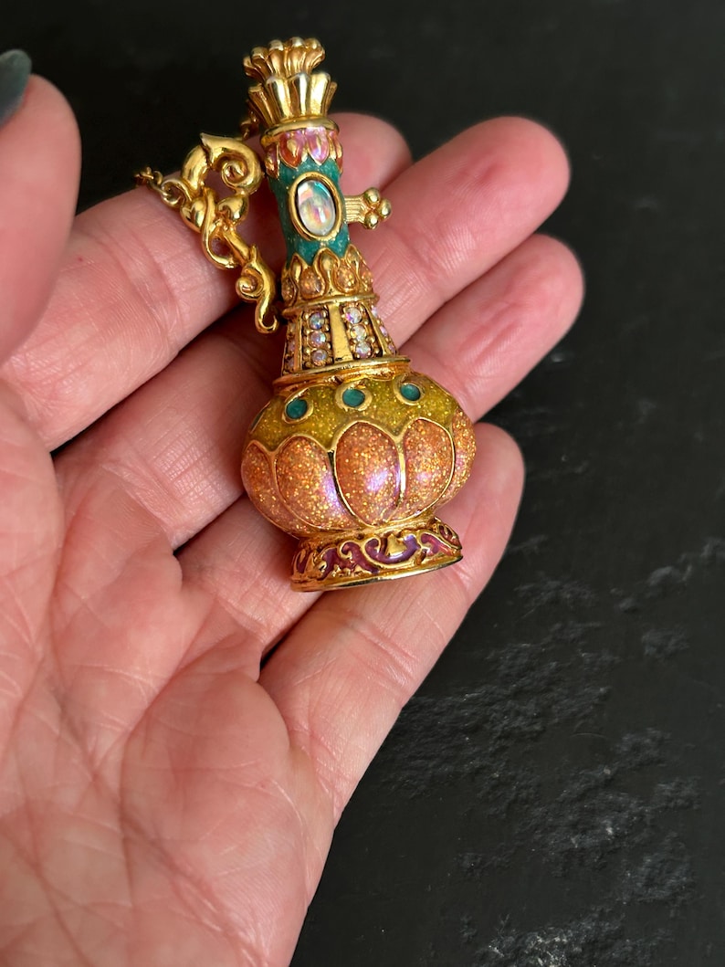 May include: A gold-toned, ornate perfume bottle pendant with a bulbous body and a slender neck. The bottle features a glittery orange and yellow body, teal accents, and a central oval-shaped gem. The pendant is embellished with small crystals and intricate gold detailing.