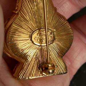 May include: Close-up of a gold-toned brooch with a pin closure. The brooch has a textured, sunburst design on the back. The brooch has an oval-shaped emblem with an inscription. The brooch is held in a person's hand.