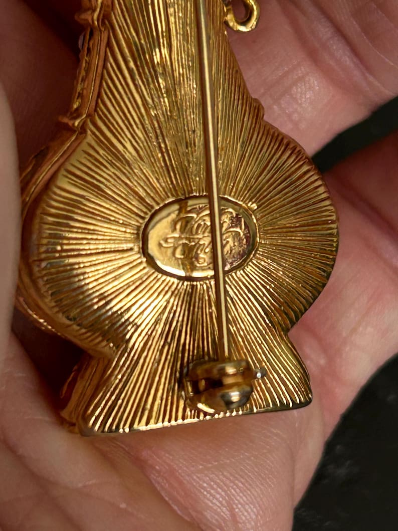 May include: Close-up of a gold-toned brooch with a pin closure. The brooch has a textured, sunburst design on the back. The brooch has an oval-shaped emblem with an inscription. The brooch is held in a person's hand.