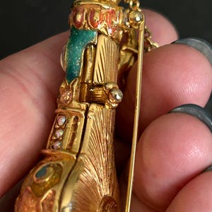 May include: A gold-toned vintage brooch in the shape of a musical instrument, possibly an accordion or a similar wind instrument. The brooch features intricate detailing, including colorful enamel accents in teal, red, and orange, and small, sparkling gemstones.