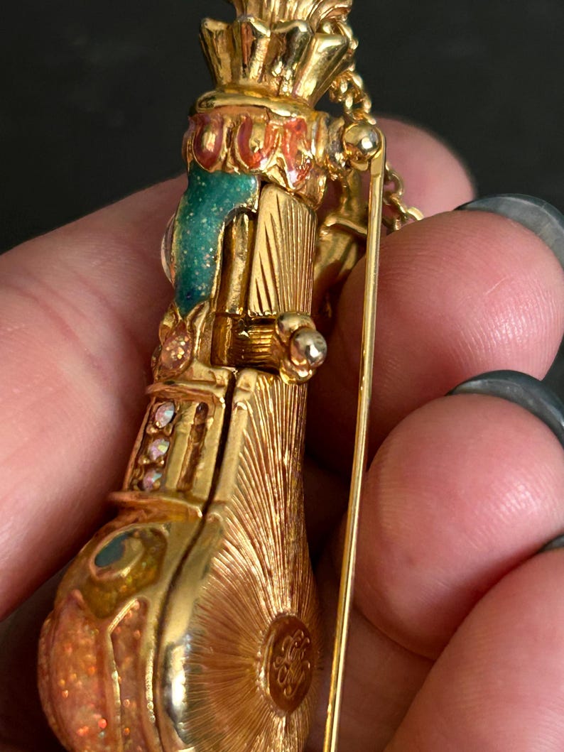 May include: A gold-toned vintage brooch in the shape of a musical instrument, possibly an accordion or a similar wind instrument. The brooch features intricate detailing, including colorful enamel accents in teal, red, and orange, and small, sparkling gemstones.