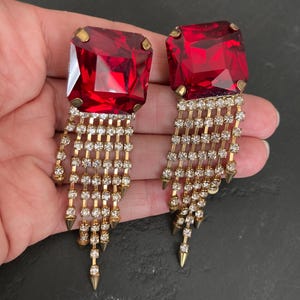 May include: A pair of statement earrings featuring large, square-cut red glass stones set in gold-tone metal. Each earring has a cascading fringe of multiple gold-tone chains adorned with small clear rhinestones, ending in pointed metal spikes.