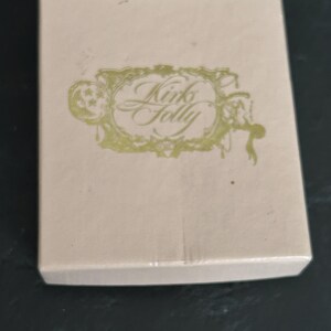 May include: A rectangular, light beige box with the words "Kinks Folly" in an ornate, gold-colored script. The text is within a decorative, gold-colored frame. The box is on a dark surface.