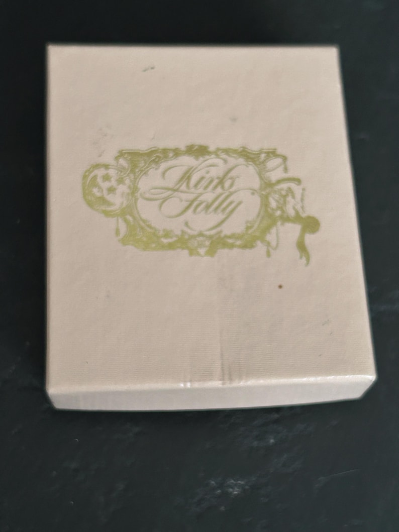 May include: A rectangular, light beige box with the words "Kinks Folly" in an ornate, gold-colored script. The text is within a decorative, gold-colored frame. The box is on a dark surface.