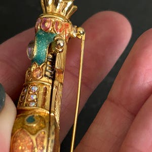 May include: A gold-toned brooch with intricate detailing. The brooch features a cylindrical design with a crown-like top and is adorned with colorful enamel and iridescent accents. A long pin extends from the back for secure fastening.