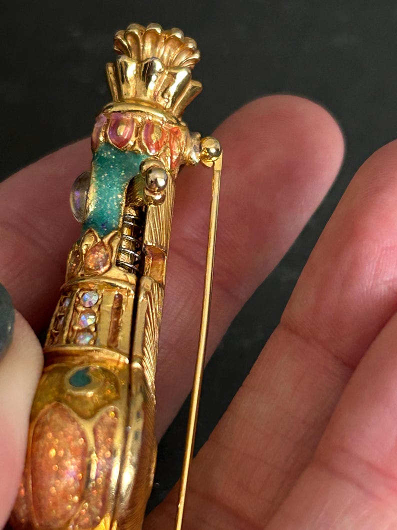 May include: A gold-toned brooch with intricate detailing. The brooch features a cylindrical design with a crown-like top and is adorned with colorful enamel and iridescent accents. A long pin extends from the back for secure fastening.