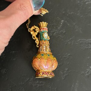 May include: A detailed, ornate gold-colored perfume bottle with a matching stopper. The bottle features a colorful design with orange, green, and red enamel, and is embellished with small, sparkling crystals. A delicate gold chain is attached.