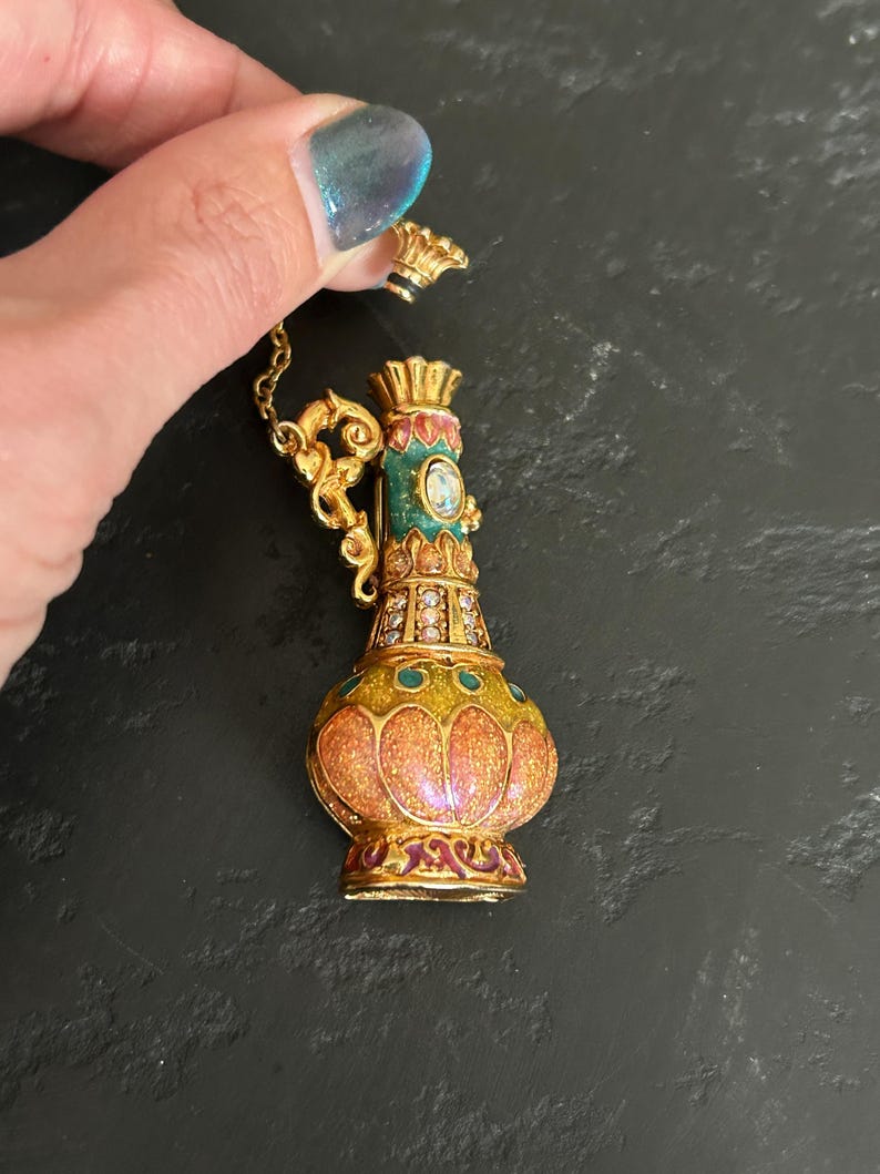 May include: A detailed, ornate gold-colored perfume bottle with a matching stopper. The bottle features a colorful design with orange, green, and red enamel, and is embellished with small, sparkling crystals. A delicate gold chain is attached.