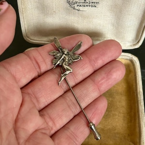 May include: A silver-toned metal stick pin featuring a detailed fairy figure with outstretched wings at the top and a cylindrical end cap. The pin is presented in a cream-colored jewelry box with the text M. Hunter Joy Goldsmith 9 Victoria Street Paignton.