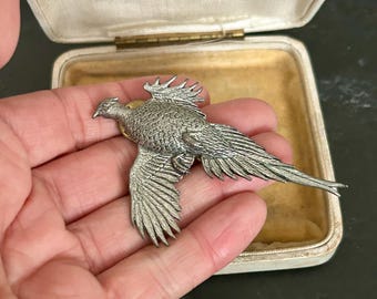 May include: A silver-toned metal brooch shaped like a flying pheasant with detailed feather textures. The brooch is presented in a rectangular, off-white hinged jewelry box with a cream-colored interior lining and a printed label on the lid.