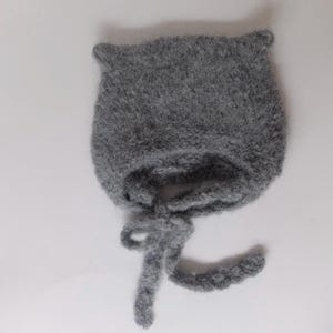 May include: A grey knitted baby bonnet with a long tie. The bonnet is made of a soft, fuzzy yarn.