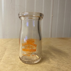 May include: A clear glass milk bottle with a rounded top and a narrow neck. The bottle has orange lettering that reads "Zenda Farms Golden Guernsey" and "Clifton, N.Y." The bottle is empty and sits on a light brown surface.