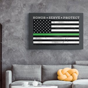 May include: A framed canvas print depicting a distressed American flag with a green stripe. The text "HONOR * SERVE * PROTECT" is above the flag. The artwork is displayed on a grey wall above a grey sofa.