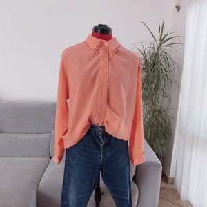 Può includere: Una blusa a maniche lunghe color pesca con un colletto in pizzo e chiusura con bottoni. La blusa è indossata sopra un paio di jeans blu.