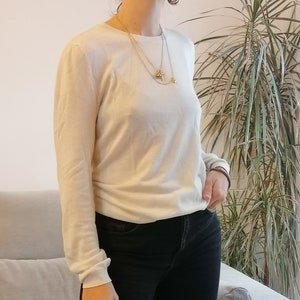 Puede incluir: Un jersey crema de manga larga y unos vaqueros negros. El jersey tiene cuello redondo y corte holgado. Se ve un collar dorado con colgantes. El conjunto es sencillo e informal, adecuado para el uso diario.
