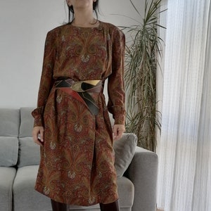 May include: A long-sleeved paisley print dress with a brown leather belt. The dress has a round neckline and a slightly flared skirt. The model is wearing brown knee-high boots.