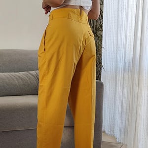 May include: Mustard yellow high-waisted pants with a wide leg. The pants have a tailored fit with pockets and a button closure. The model is wearing gold-colored heeled sandals. The pants are made of a lightweight fabric.