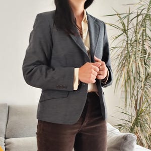Könnte beinhalten: Ein anthrazitfarbener Blazer mit drei Knöpfen am Ärmel, kombiniert mit einer hellen Bluse und einer braunen Cordhose. Das Outfit ist ein klassischer Business-Casual-Stil, geeignet für ein professionelles Umfeld.