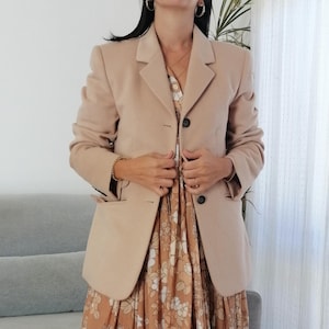 Puede incluir: Una chaqueta beige con cierre de dos botones, puesta sobre un vestido largo floral con estampado marrón y blanco.