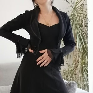 May include: A black dress with a ruffled hem and a matching black jacket with ruffled sleeves. The jacket has a shawl collar and a single button fastening.