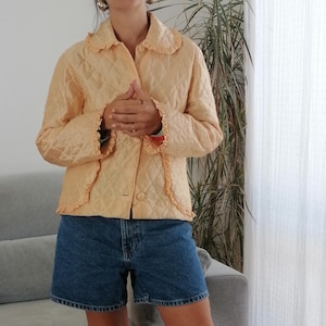 Könnte beinhalten: Eine peachfarbene gesteppte Jacke mit Rüschenkragen und -ärmeln. Die Jacke hat einen Knopfverschluss und ein rautenförmiges Steppmuster.