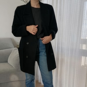 Puede incluir: Un abrigo negro de lana de doble botonadura con un corte largo y relajado. El abrigo tiene bolsillos grandes y se lleva sobre una camiseta gris y unos vaqueros azules.