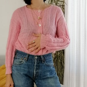 May include: A pink knitted cardigan with a single button closure. The cardigan has a slightly oversized fit and long sleeves. The model is wearing the cardigan with blue jeans.