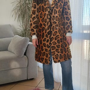 Könnte beinhalten: Ein langer Mantel mit Leopardenmuster in Braun und Schwarz, mit doppelreihigem Design. Der Mantel hat einen breiten Kragen und Knöpfe. Die Person trägt eine blaue Jeans und orangefarbene Turnschuhe.