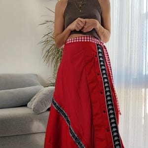 以下が含まれることがあります： 黒と白のストライプがサイドに施された赤いラップスカート。スカートはギンガムチェックのウエストバンドとボタン留め。軽量な生地で作られており、暖かい天候に最適です。