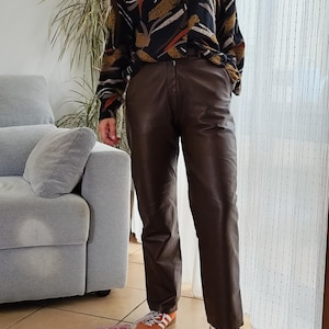Pode incluir: Uma pessoa vestindo uma camisa preta com um padrão abstrato dourado, marrom e branco, combinada com calças de couro marrom e tênis laranja com listras brancas. A roupa é estilizada de forma casual.