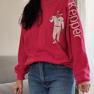 May include: A magenta sweatshirt with a baseball player graphic and the word "Keeper" printed on the sleeve. The baseball player is in a white uniform and holding a bat. The sweatshirt is worn with blue jeans.