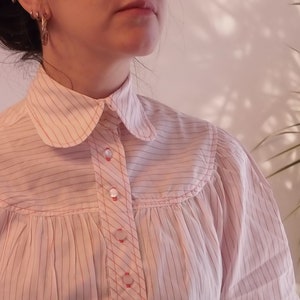 May include: A white and pink striped button-up shirt with a peter pan collar. The shirt has a gathered detail at the bust and long sleeves.
